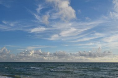 Yumuşak deniz dalgalarının kumsalda öptüğü gibi kumlu okyanus kıyısında gevşeyin. Yumuşak beyaz bulutlarla süslenmiş, dingin mavi gökyüzünün altında..