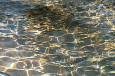 Suyun yüzeyinde canlı güneş ışığı bir çeşmede dans eder, yaşlanmış taş zeminde renkli ve soyut bir doku oluşturur.