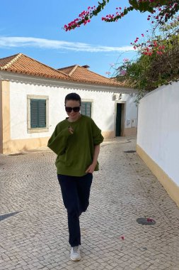 Yeşil gömlekli ve siyah güneş gözlüklü genç bir kadın eski kasabanın kaldırım taşı caddelerinde yürüyor..