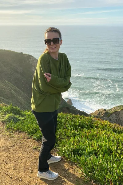 Young woman in green jacket and sunglasses standing on a cliff and looking at the sea