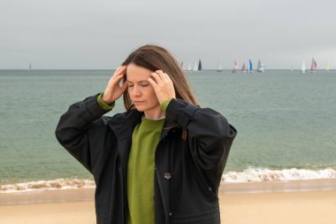 Genç bir kadın sahilde dikilip başını çevirirken baş ağrısı çekiyor.