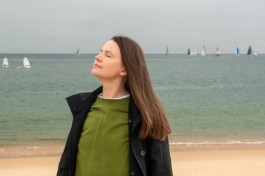 Genç bir kadın denizin önündeki sahilde dinleniyor ve yelkenli sürüyor.