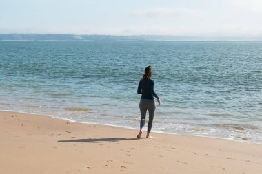 Güzel genç bir kadın sahilde yürüyor, denize bakıyor.