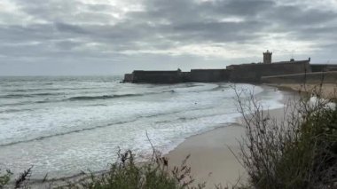 Sandy Praia da Torre plajı, bulutlu arka planda. Sao Juliao da Barra Ortaçağ Kalesi. Dramatik deniz manzarası ve Atlantik Okyanusu kıyısı.