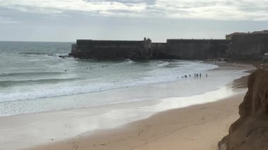 Sandy Praia da Torre plajı, bulutlu arka planda. Sao Juliao da Barra Ortaçağ Kalesi. Dramatik deniz manzarası ve Atlantik Okyanusu kıyısı.