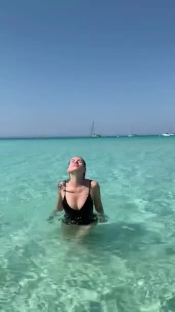 一个身穿黑色泳衣的年轻女子从清澈的蓝水中出现在一个空旷的海滩上 — 图库视频影像