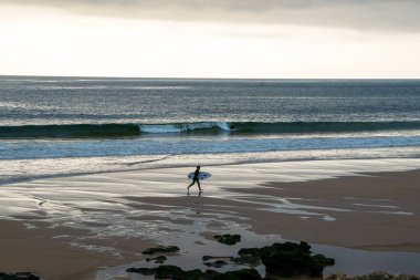 Gün batımında sahilde yürüyen bir sörfçünün silüeti. Oeiras, Portekiz, 9 Şubat 2023