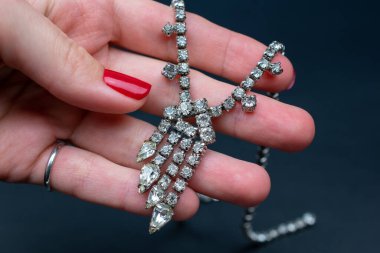 Kırmızı manikürlü bir el siyah arka planda elmas bir kolye tutuyor.