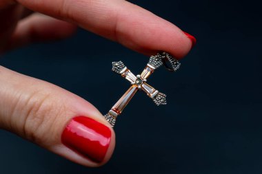 Siyah arka planda gümüş haç tutan kırmızı manikürlü bir kadın eli.