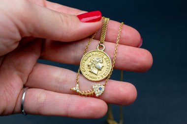 Kırmızı manikürlü bir kadın, siyah arka planda altın bir madalyon taşıyor.