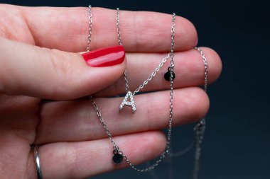 Kırmızı manikürlü bir kadın, koyu arkaplanda gümüş bir kolye tutuyor.