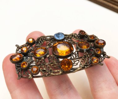 Beyaz zemin üzerinde renkli taşlar olan metal bir broşu tutan el.