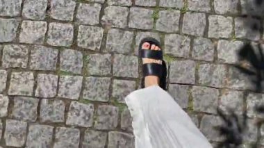 Siyah sandaletli kadın bacaklarının kaldırım taşları boyunca yürüyüşünün üst görüntüsü.