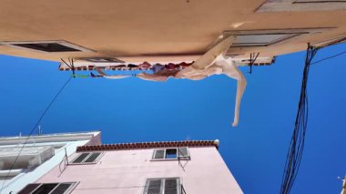 Upward view of building facades with laundry hanging on clotheslines against blue sky, windy day atmosphere