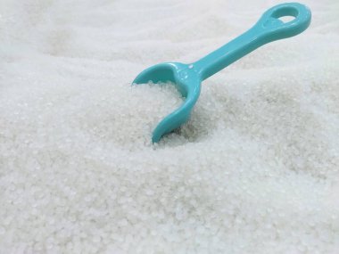 Yakın plan çekimde, beyaz kumdan bir yatakta dinlenen açık mavi plastik bir oyuncak kum kepçesi yer alır.