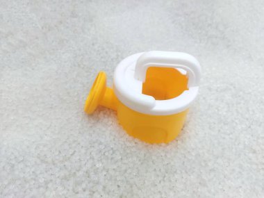 Küçük, sarı plastik bir oyuncak sulama kutusu, beyaz saplı ve balçıklı, yapay beyaz kumdan bir yatakta dinleniyor.