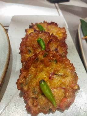 Yakın çekimde üç altın kahverengi Endonezyalı Bakwan Jagung (mısır böreği) dikdörtgen açık gri bir tabakta dizilmiş.