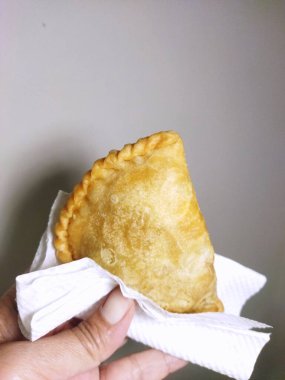 Bir el, beyaz kağıt peçetenin üzerinde duran kıvrımlı kenarlı, tek bir altın-kahverengi, kızarmış körili puf veya pastel tutuyordu.