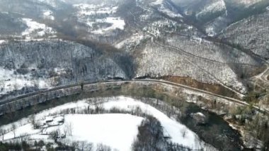 Dağların eteklerinde bir yol esiyor, kış manzarasında bir nehrin kıyısından geçiyor.