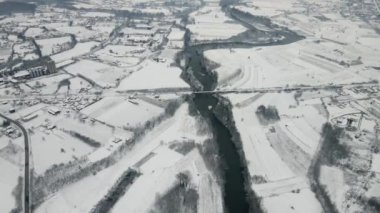 Bir hava manzarası, karla kaplı tarlalar ve kış manzarasındaki bir kasabayı saran bir nehri yakalar.
