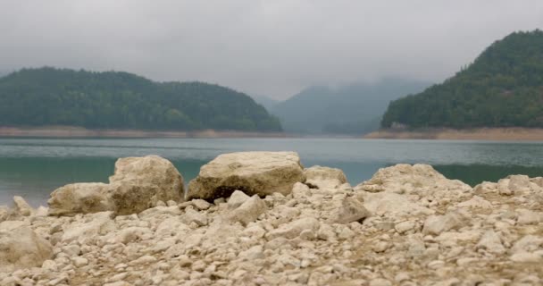 セルビアのザイツァーンの背景にある霧の中の丘を持つ湖岸の大きな岩 — ストック動画