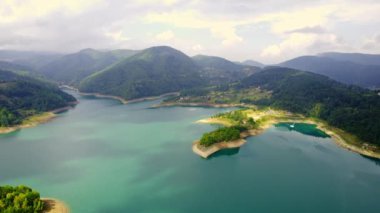 İnsansız hava aracı bulutlu havada ağaçlık tepelerle çevrili açık mavi fırına iniyor.