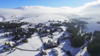 İnsansız hava aracı bulutlu ve arka planda karla dağlardaki küçük turistik köye doğru uçuyor.