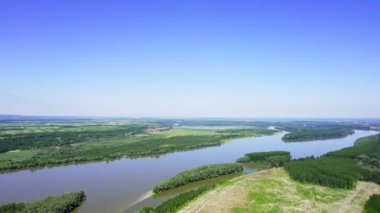 İnsansız hava aracı Tuna Nehri 'nin üzerinde uçar ve onu çevreleyen yemyeşil araziyi parlak mavi gökyüzünün arka planına karşı yakalar.