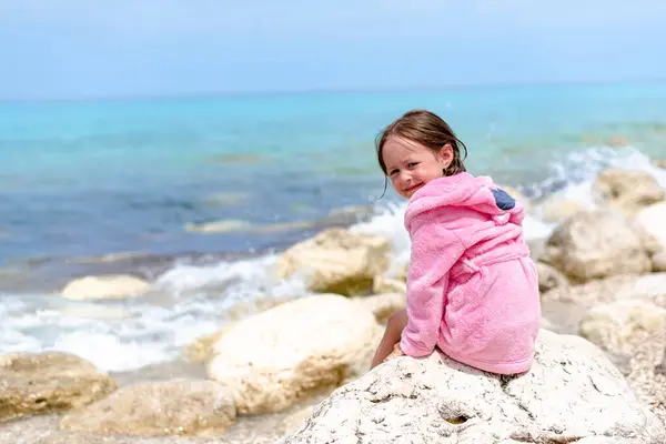 Pembe bornozlu küçük bir kız su kenarında kameraya bakarak eğleniyor.