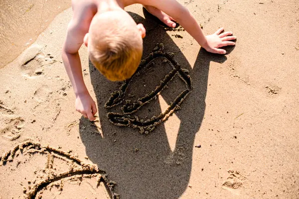 Kumsalda kumlara resim çizen küçük bir sarışının üst açı görüntüsü.