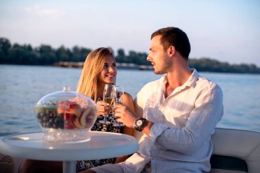Genç bir çift teknede eğleniyor, şampanya içiyor ve gülümsüyor.