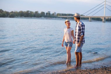 Çiftler, arkalarında köprü olan sığ nehir sularında yürüyorlar.