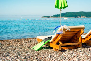 Çakıl taşı plajı cenneti, dalgıç maskesi, yüzgeçler ve şnorkel tahta sandalyede.