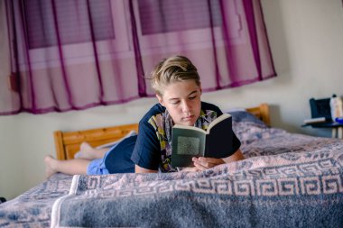 Genç çocuk yatağın üstünde kitap okuyor.