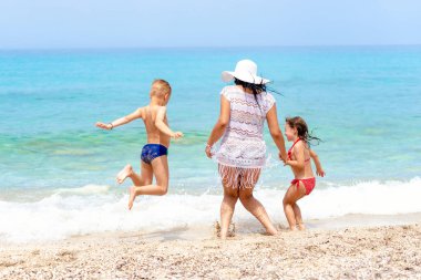 Anne ve çocuklar turkuaz suyun kıyısında eğlenip zıplıyorlar.