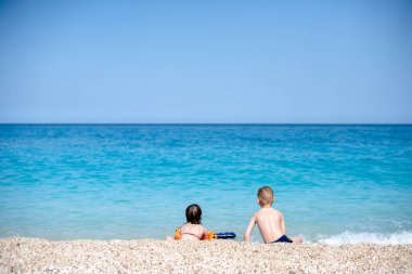 Küçük bir kız ve oğlan güzel, çakıllı sahilde suda oynuyorlar.