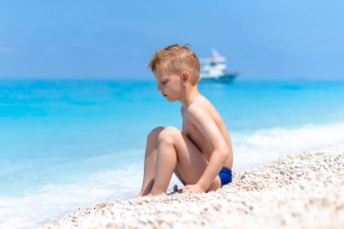 Bir çocuk turkuaz suyun kıyısındaki güzel, çakıllı sahilde oynuyor.