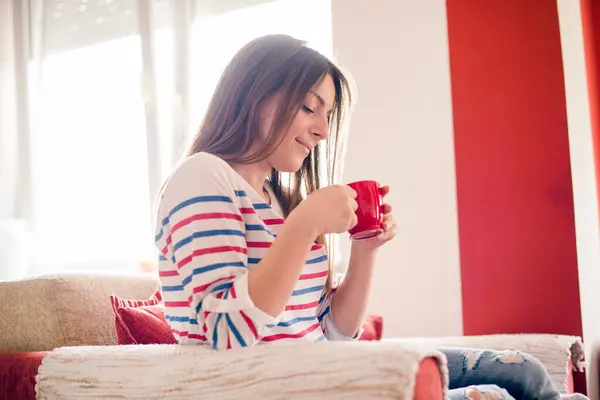 Oturma odasındaki kanepede kahve içen genç kadın.