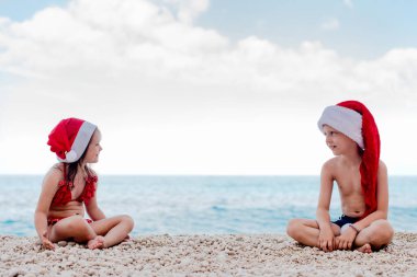 Noel Baba şapkalı küçük kız ve oğlan suyun kenarında oturuyor. Modern Noel kutlaması