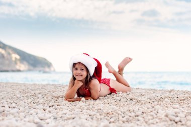 Güzel çakıl taşı kumsalında Noel Baba şapkalı mutlu küçük kız. Kameraya bakıyor.