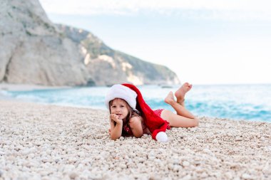 Güzel çakıl taşı kumsalında Noel Baba şapkalı mutlu küçük kız. Kameraya bakıyor.