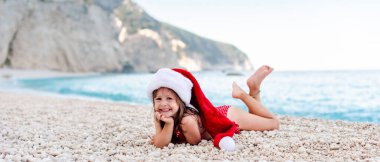 Güzel çakıl taşı kumsalında Noel Baba şapkalı mutlu küçük kız. Kameraya bakıyor.