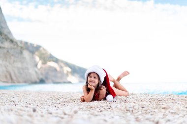 Güzel çakıl taşı kumsalında Noel Baba şapkalı mutlu küçük kız. Kameraya bakıyor.