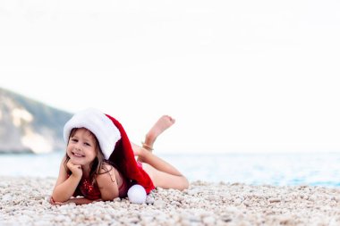 Güzel çakıl taşı kumsalında Noel Baba şapkalı mutlu küçük kız. Kameraya bakıyor.