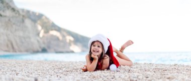 Güzel çakıl taşı kumsalında Noel Baba şapkalı mutlu küçük kız. Kameraya bakıyor.