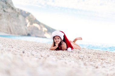 Güzel çakıl taşı plajında Noel Baba şapkalı mutlu küçük kız. Modern Noel kutlaması.