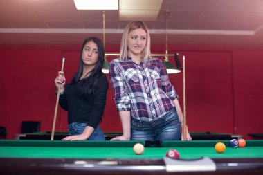 İki çekici kız, sarışın ve esmer, bilardo masasının yanında poz veriyorlar.