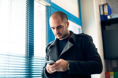 Ofiste cep telefonuyla yazan genç girişimci, pencerenin yanında duruyor..