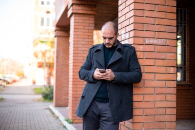 Beyaz iş adamı cep telefonuyla mesajlaşıyor, sokak duvarına yaslanıyor..