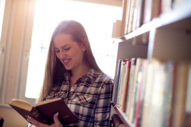 Kütüphanede gülümseyen genç kız, kitaplığın yanında duruyor ve kitap okuyor..